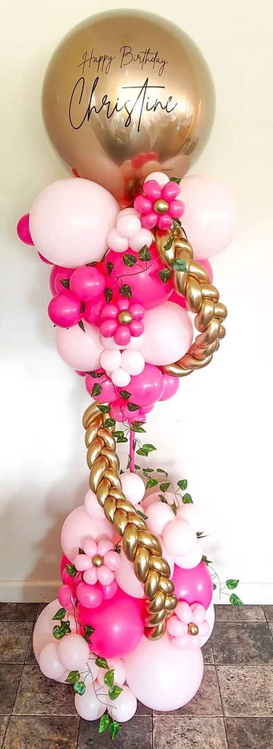 Pink and fuchsia balloon column with balloon flowers, greenery and a gold braided twist, topped with a gold bubble balloon reading Happy Birthday Christine
