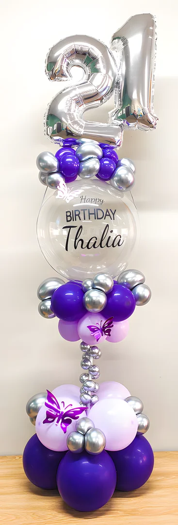 Silver 21 foil numbers above a clear bubble balloon reading Happy Birthday Thalia, with butterfly accents on a purple and lilac balloon column