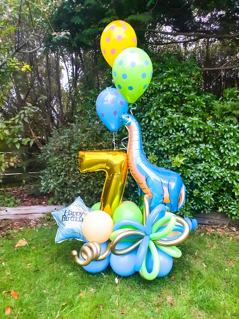 Gold 7 foil number with a blue dinosaur foil balloon, polka dot helium balloons and a blue and green twisted balloon base in a garden