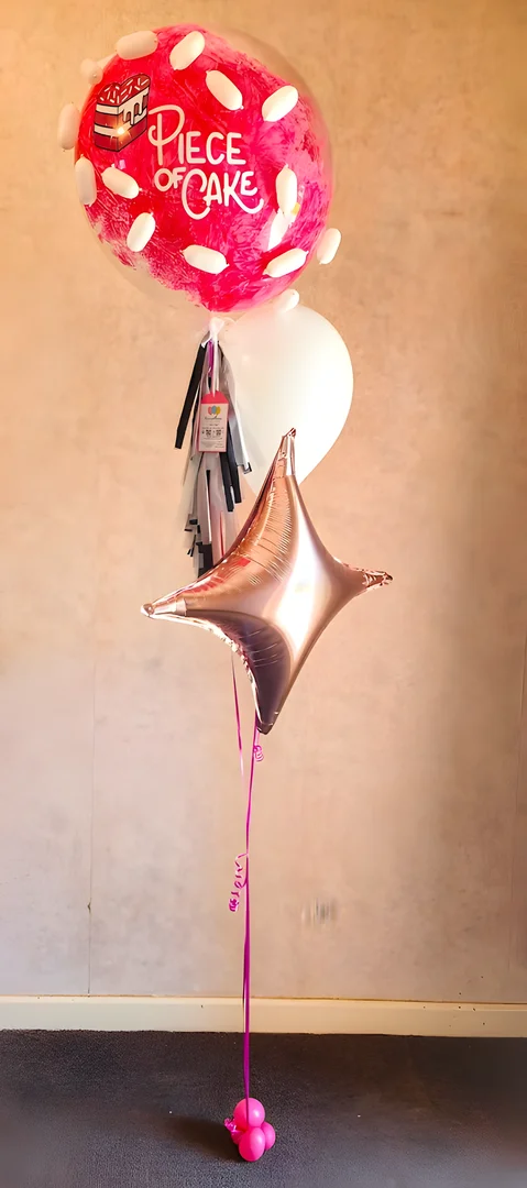 Pink Piece of Cake bubble balloon with black and silver tassels, a white balloon and a rose gold foil star, on a pink balloon weight