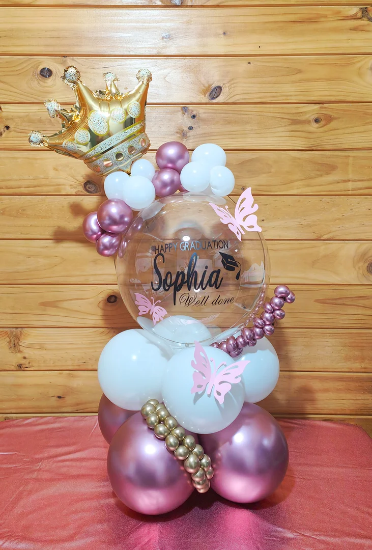 Clear graduation bubble balloon reading Happy Graduation Sophia with a gold crown balloon, pink butterflies and white and mauve balloons