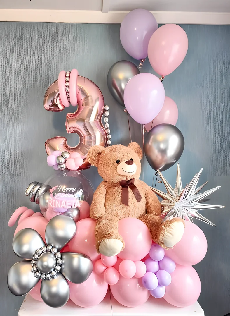 Large teddy bear sitting on a pink balloon base with a rose gold 3 foil number, a personalised bubble balloon, a silver starburst and pink and lilac helium balloons