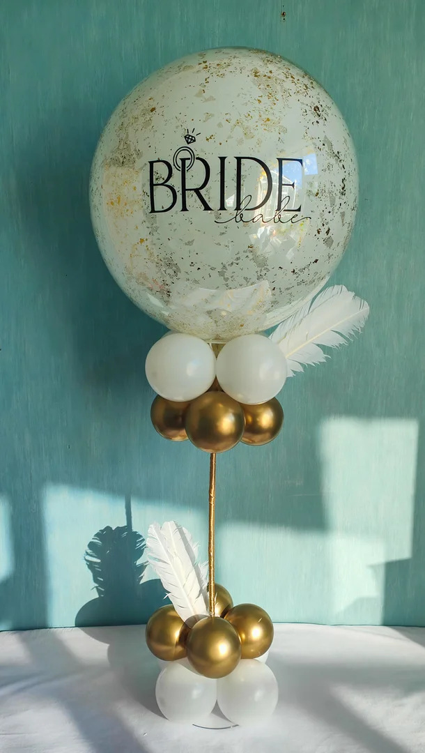 Clear bubble balloon reading Bride Babe with shimmering gold foil inside, white feathers and white and gold balloons on a balloon stand